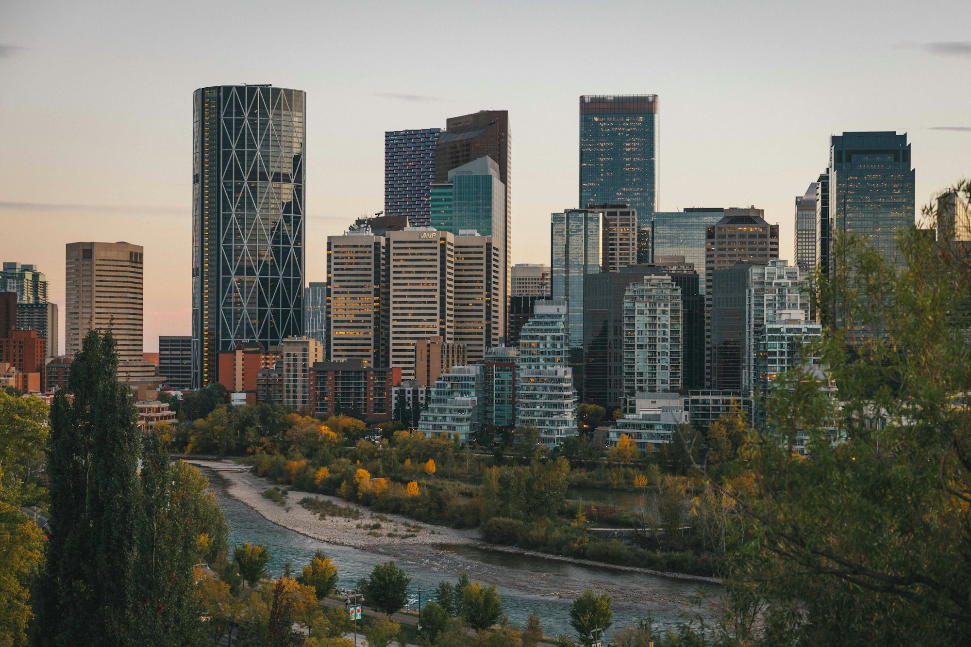 Downtown Calgary office towers above the Bow River in early autumn, with yellowing trees along the riverbank.